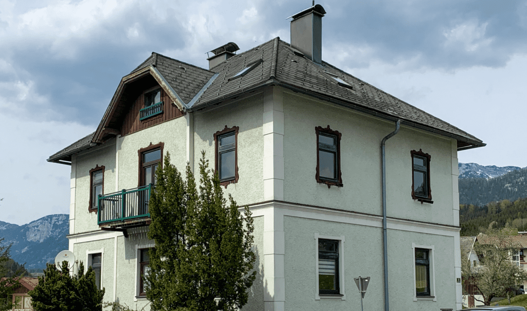 Ein altes Haus in Österreich mit grüner Fassade und weißen Ziersteinen vor einem gebirgigen Hintergrund